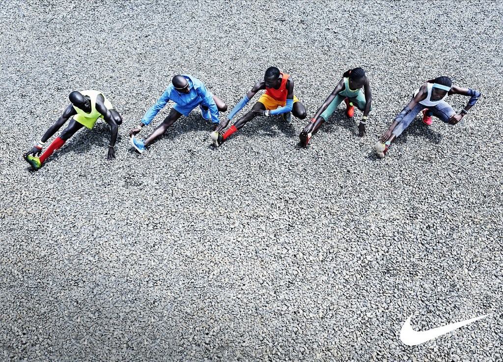 Overhead shot of five runners in sportswear stretching outdoors on a gravel surface with the Nike logo in the lower corner.