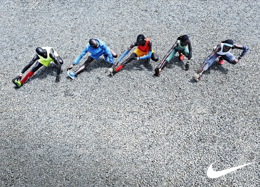 Overhead shot of five runners in sportswear stretching outdoors on a gravel surface with the Nike logo in the lower corner.