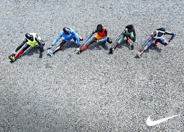 Overhead shot of five runners in sportswear stretching outdoors on a gravel surface with the Nike logo in the lower corner.