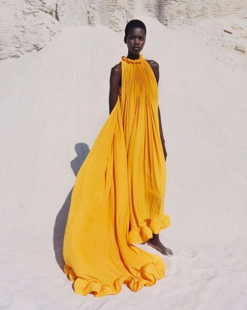 Woman in flowing yellow gown stands on sand dune in desert landscape, bathed in warm light, showcasing fashion and nature.