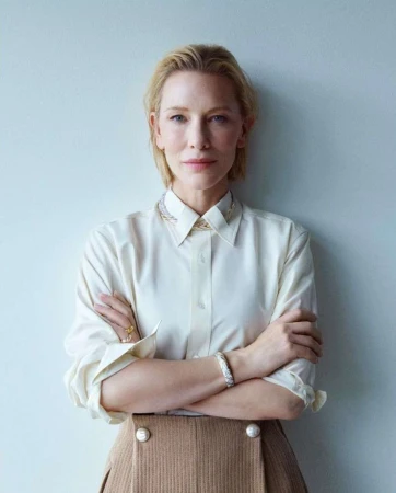 Portrait of Cate Blanchett with arms crossed wearing a white button-down shirt and brown skirt against a neutral wall