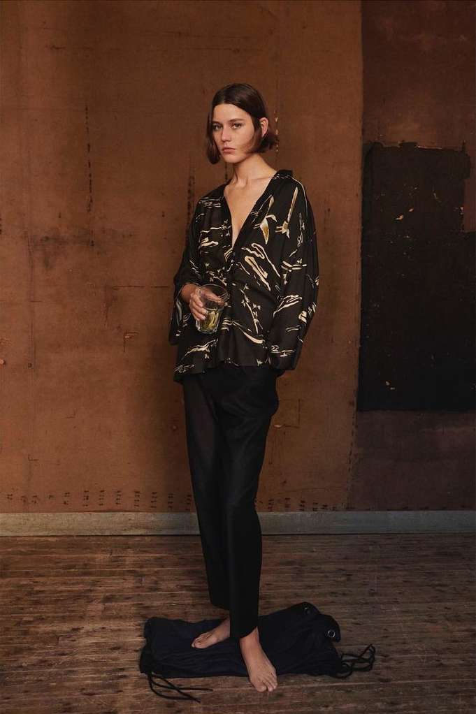 A young woman with short brown hair stands in a rustic indoor setting wearing a patterned blouse and dark trousers, holding a glass.