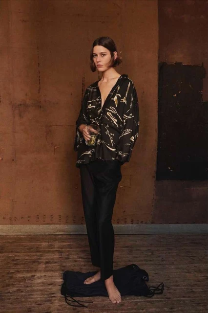 A young woman with short brown hair stands in a rustic indoor setting wearing a patterned blouse and dark trousers, holding a glass.