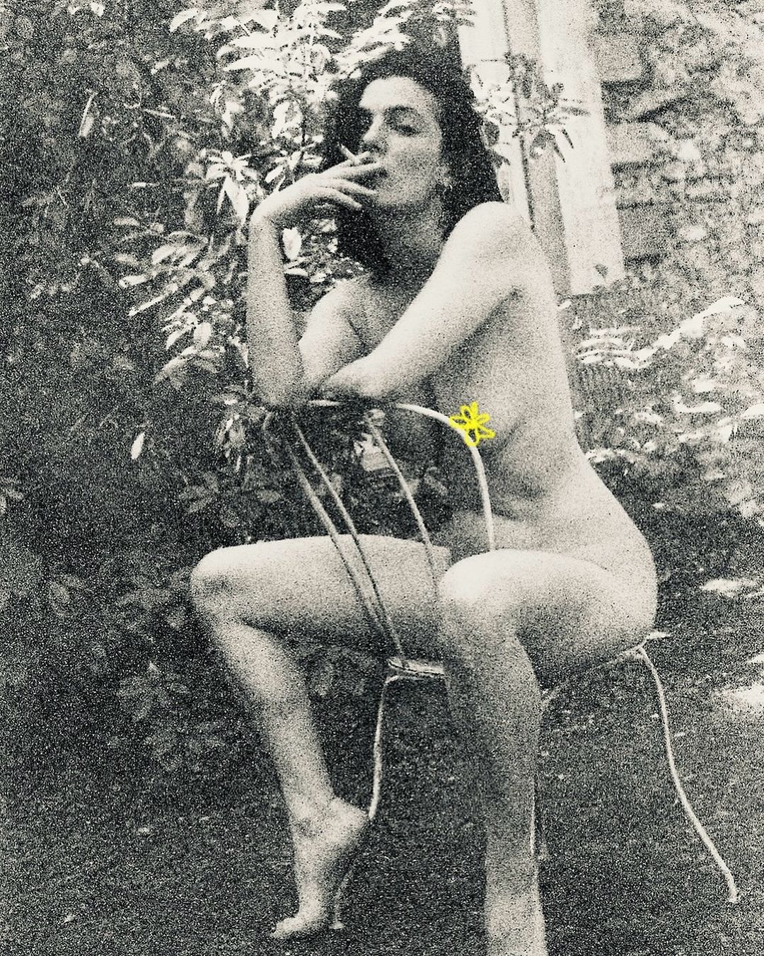 Monochrome portrait of a nude woman smoking, sitting on a metal chair outdoors with foliage in the background, creating a vintage aesthetic.