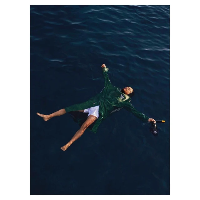 Adult male with dark hair floats on his back in ocean wearing green track jacket, white shorts, and holding a dark object in his hand.