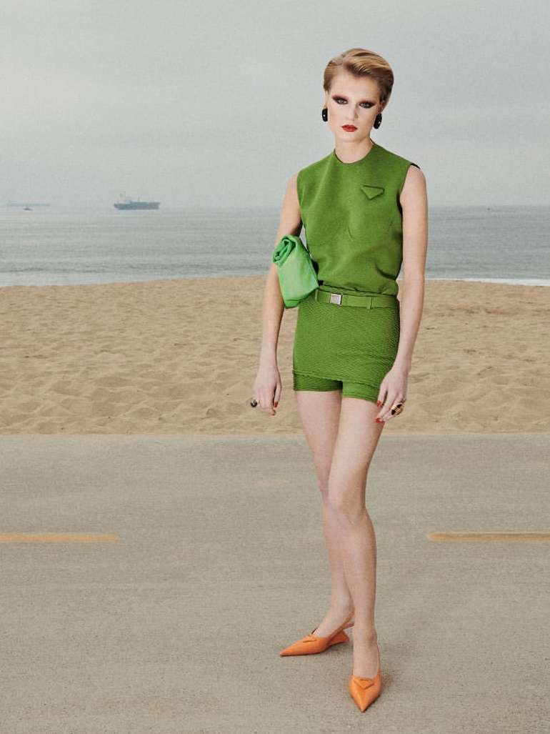 Woman in green outfit poses on beachside road with ocean backdrop, showcasing fashion and beachside style in editorial photography.