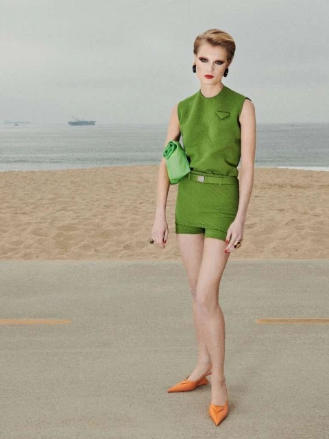 Woman in green outfit poses on beachside road with ocean backdrop, showcasing fashion and beachside style in editorial photography.