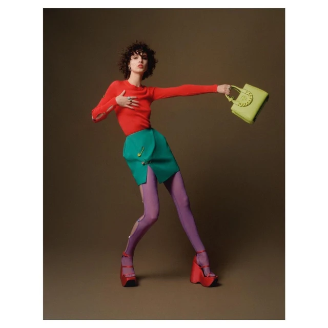 A woman poses in a red sweater, green skirt, purple tights, and platform heels, holding a yellow handbag against a brown background.