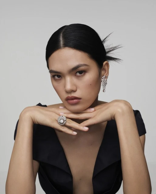 Portrait of an Asian woman with dark hair wearing a black dress, diamond ring, and earrings against a grey background in a photo studio.