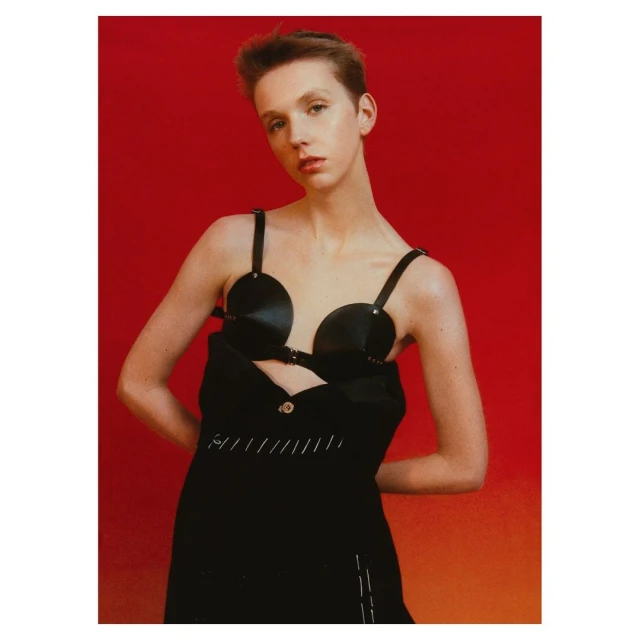 A fair-skinned person poses in a black bra-like top and black dress against a red background in a studio-lit fashion shot.