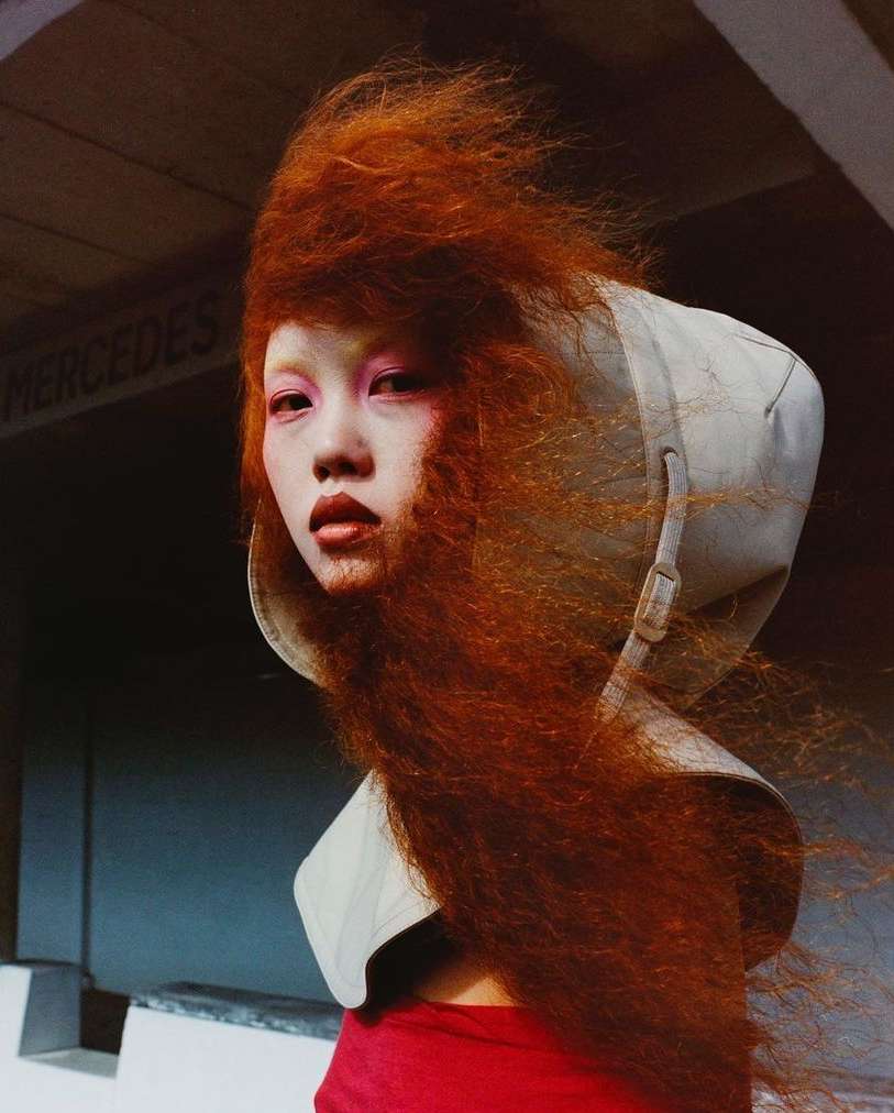 Close-up of a woman with vibrant red hair and pink makeup, wearing a high collar, set against a textured gray wall with Mercedes text.
