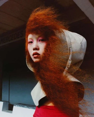 Close-up of a woman with vibrant red hair and pink makeup, wearing a high collar, set against a textured gray wall with Mercedes text.