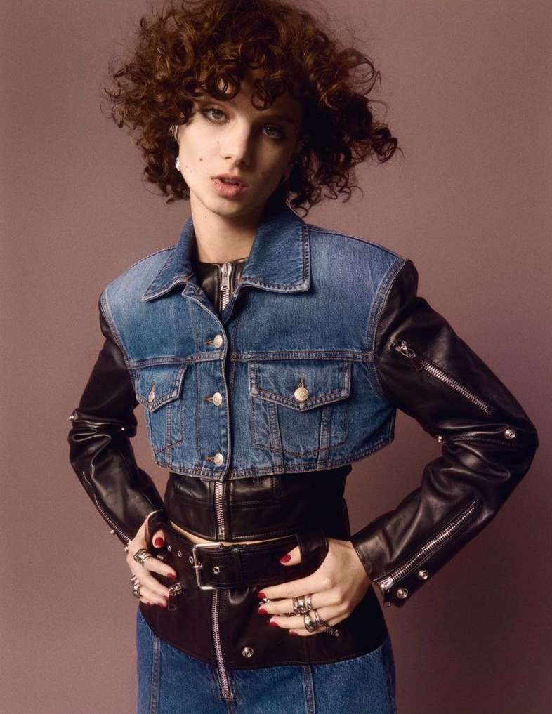 Portrait of a woman with curly hair, wearing a cropped denim jacket with leather sleeves, leather skirt and studded belt against mauve background.