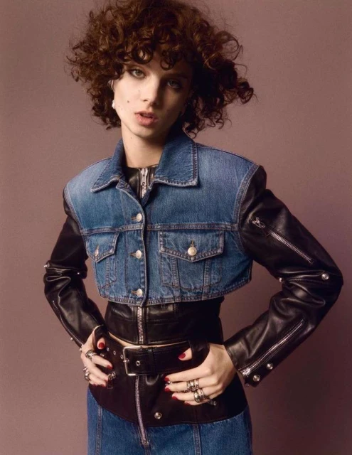 Portrait of a woman with curly hair, wearing a cropped denim jacket with leather sleeves, leather skirt and studded belt against mauve background.