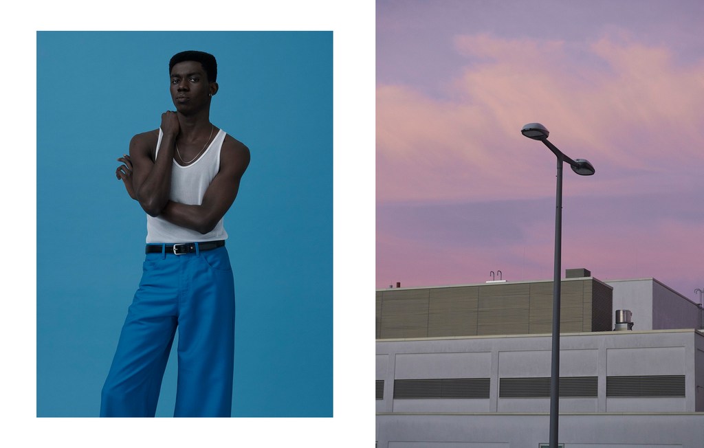 Studio portrait of a dark-skinned adult male in a white tank top and blue trousers against a solid blue backdrop; building and sky background.