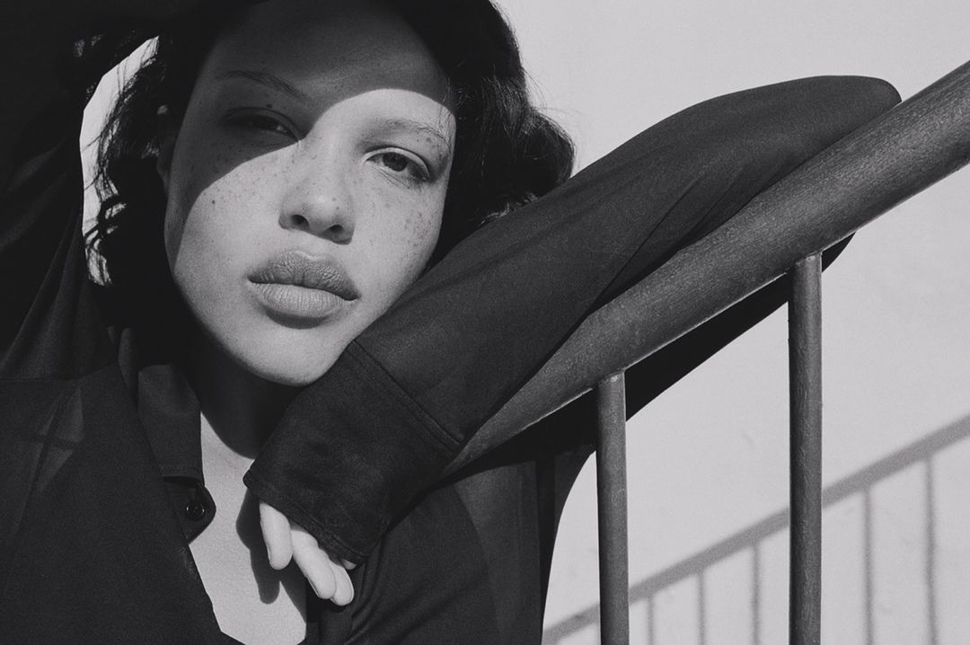 Close-up black and white portrait of a woman with freckles and a sheer top leaning against metal railing, bathed in natural sunlight.