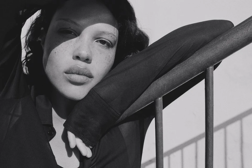 Close-up black and white portrait of a woman with freckles and a sheer top leaning against metal railing, bathed in natural sunlight.