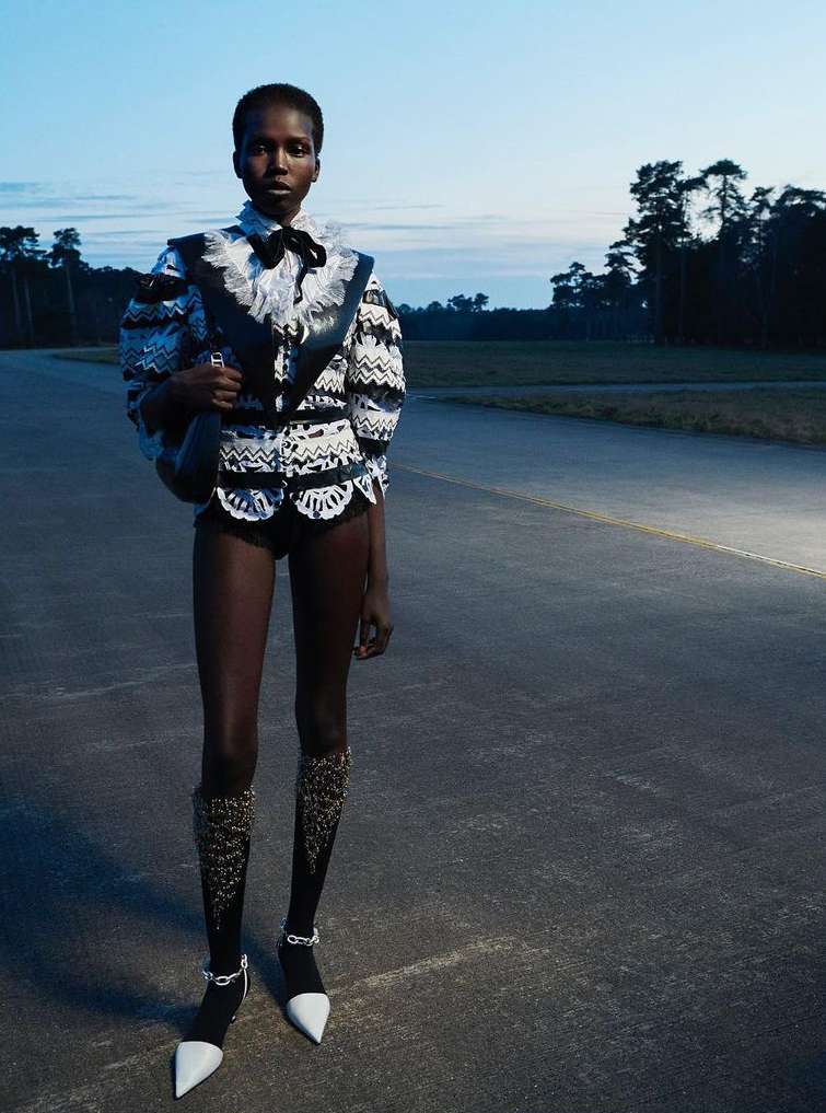 A woman in a black and white lacy jacket, shorts, and decorative knee-high socks stands on an asphalt road in an open field at dusk.