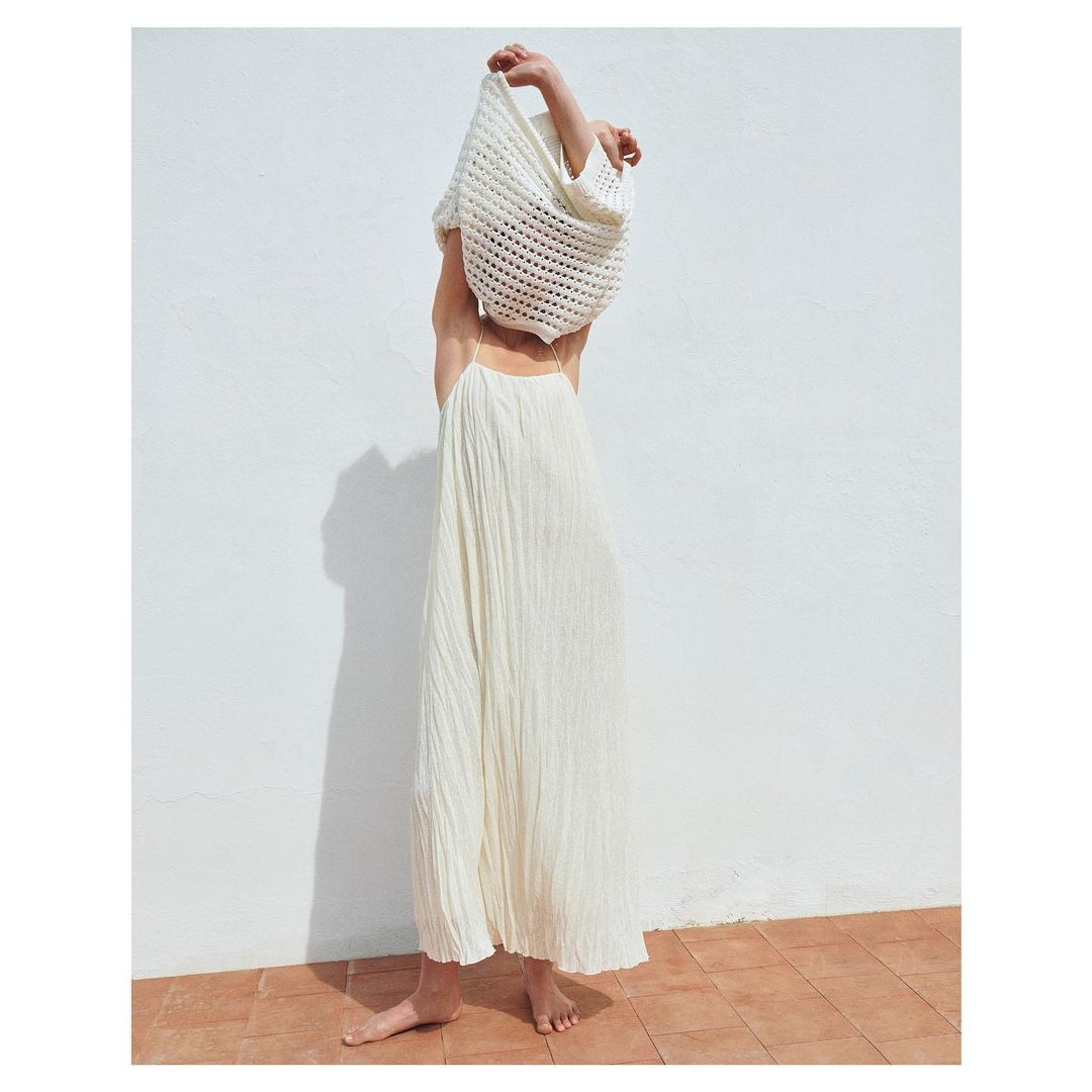 Barefoot woman in a long white dress stands near a white wall, holding up a cream-colored crochet bag over her head in an outdoor setting.