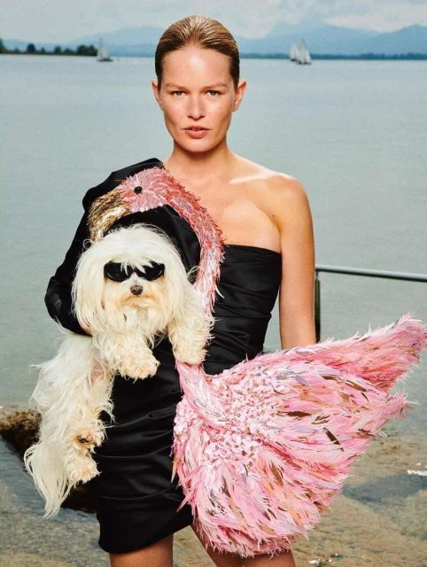 Woman in black dress holds white dog wearing sunglasses on lakeside with sailboats in background, fashion, beauty, and lifestyle.