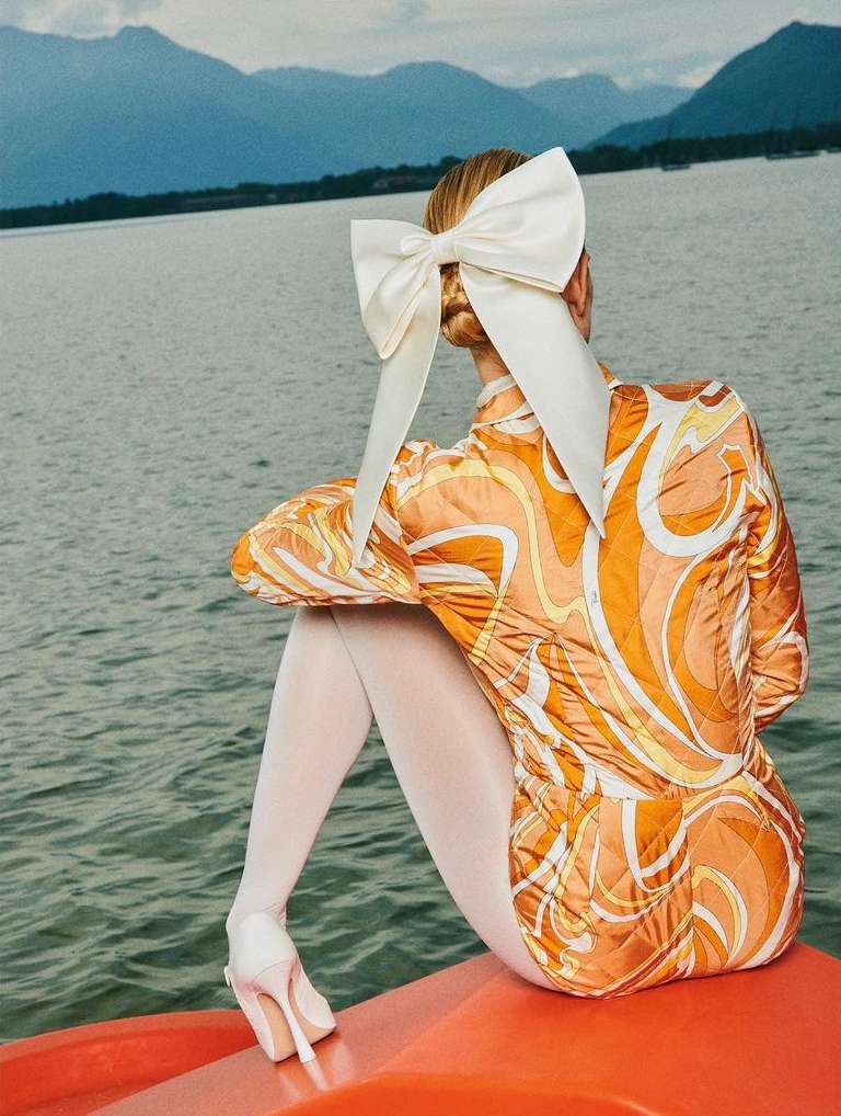 Woman seen from behind wearing an orange patterned top, white tights, and a large white hair bow by a lake with mountains.
