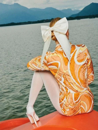 Woman seen from behind wearing an orange patterned top, white tights, and a large white hair bow by a lake with mountains.