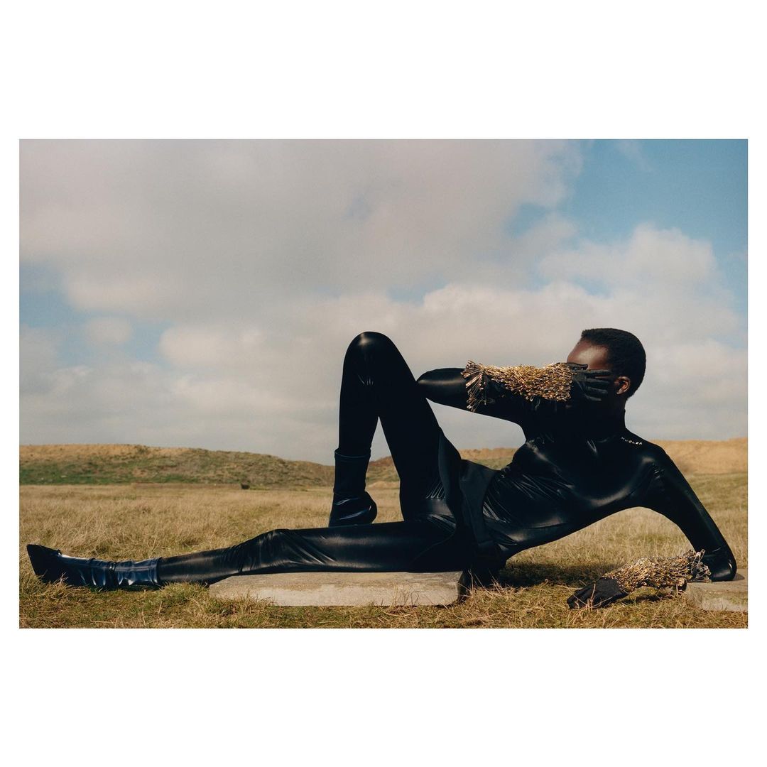 A Black model in a black catsuit and blue boots is lying down outside on a clear day with a grassy hill and cloudy skies in the background.