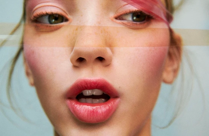 Close-up portrait of a young woman with freckles, coral lipstick, and translucent pink tape across her eyes, photographed by Julia Noni.