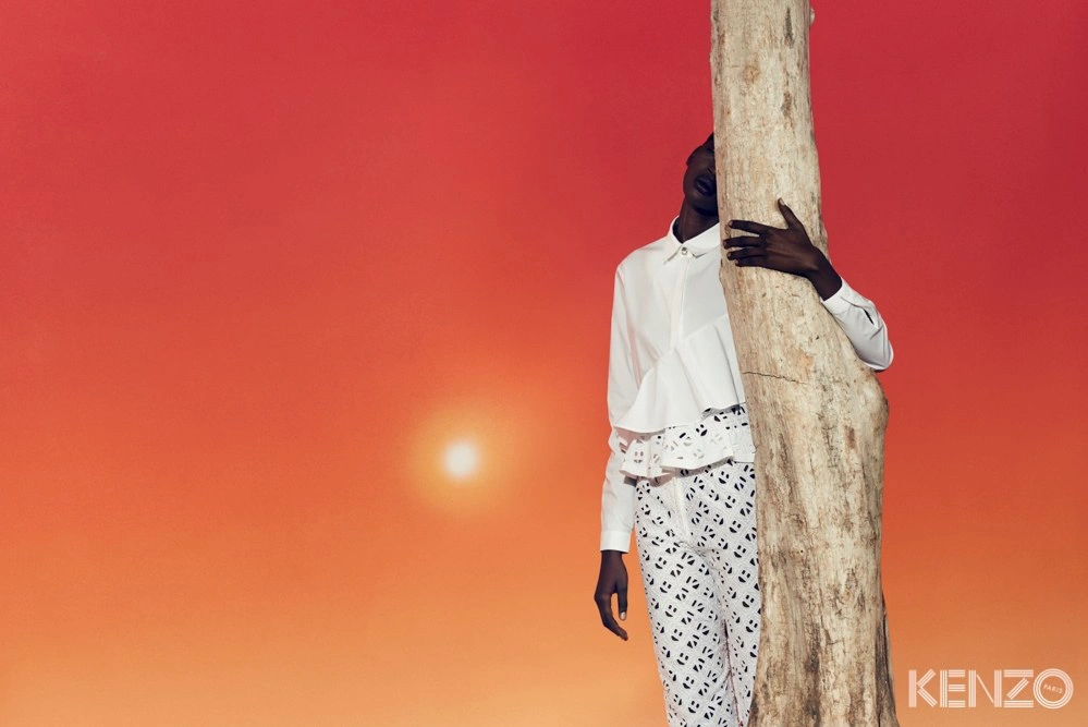 Model wearing a white shirt and patterned trousers embracing a tree trunk against a gradient orange sky with sun
