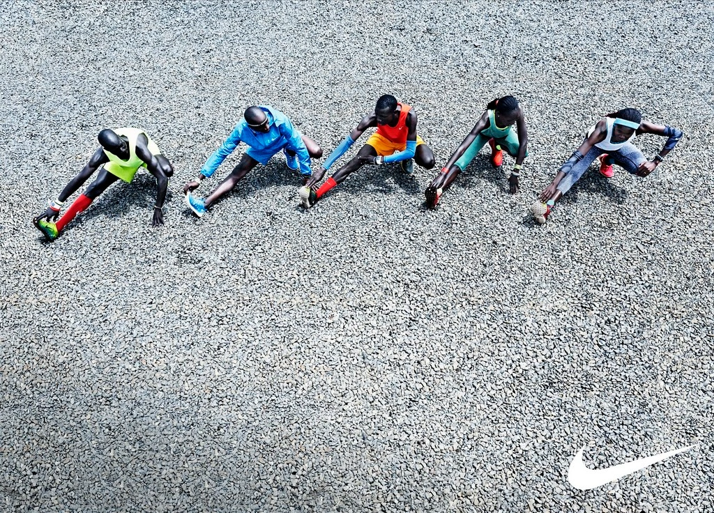 High-angle view of five runners stretching on a gravel surface wearing colourful athletic apparel.