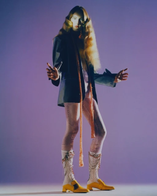 Full-length studio shot of a stylish adult woman with long blonde hair, wearing a black jacket, pink leggings, and yellow boots, posing.