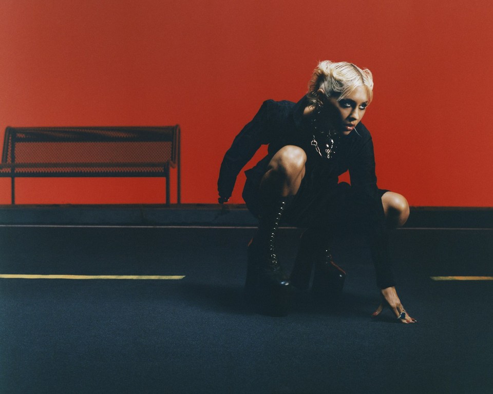 A fashion-forward, blonde adult female crouches indoors, clad in a dark jacket and platform boots, against a vivid red backdrop with bench.