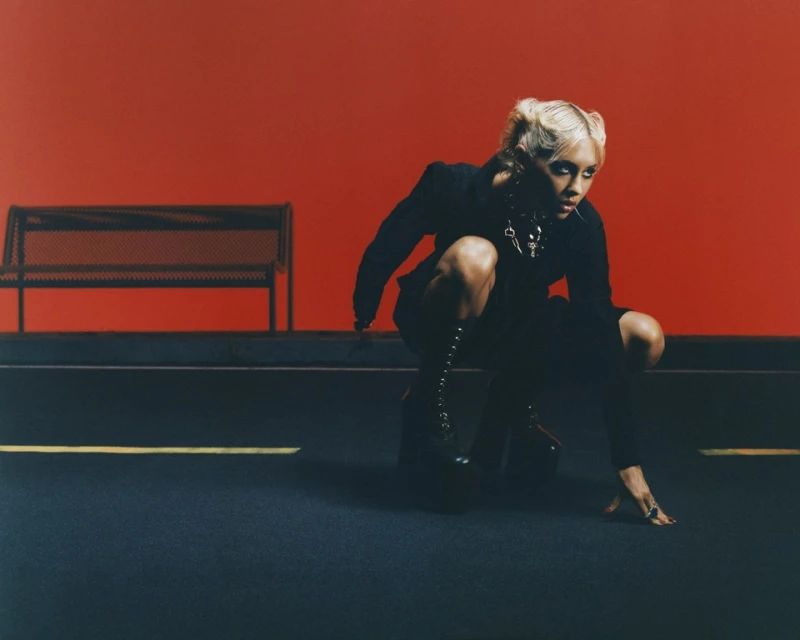 A fashion-forward, blonde adult female crouches indoors, clad in a dark jacket and platform boots, against a vivid red backdrop with bench.