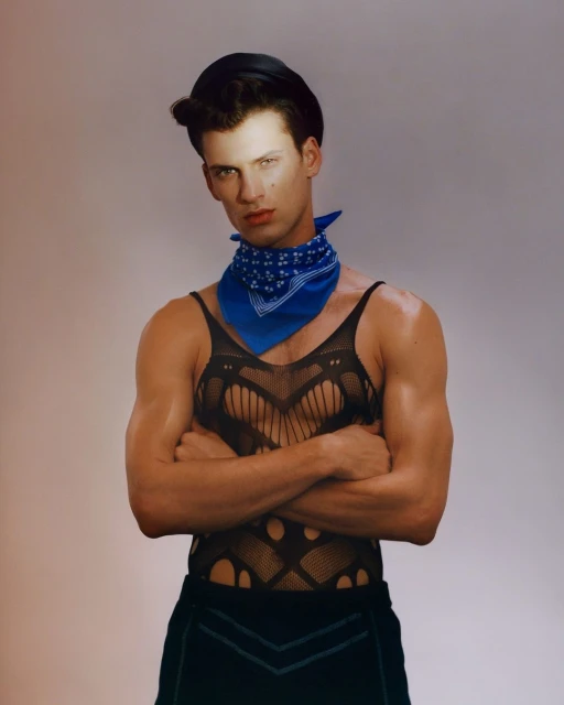 Portrait of a muscular adult male with arms crossed, wearing a black tank top, cap, and blue bandana against a neutral backdrop.