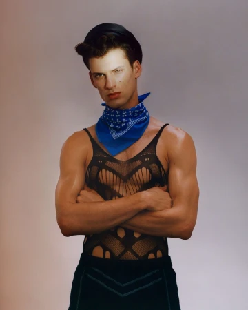 Portrait of a muscular adult male with arms crossed, wearing a black tank top, cap, and blue bandana against a neutral backdrop.
