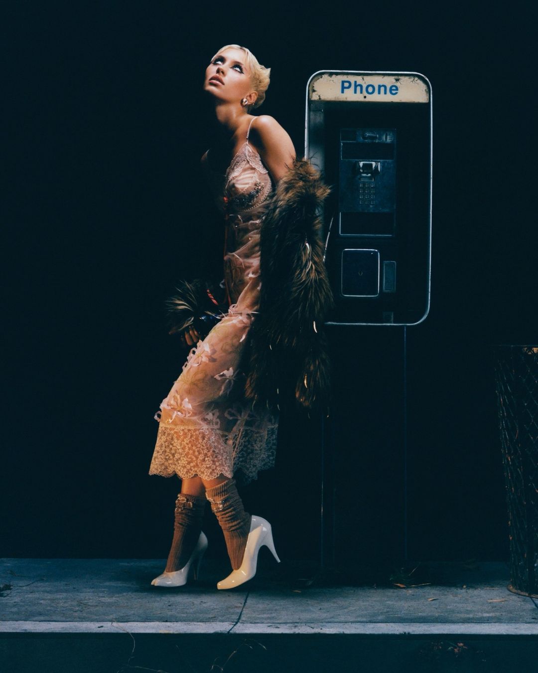 Fashion shot of a blond woman in a lace dress standing beside a vintage phone booth in a dark urban setting with dramatic lighting.