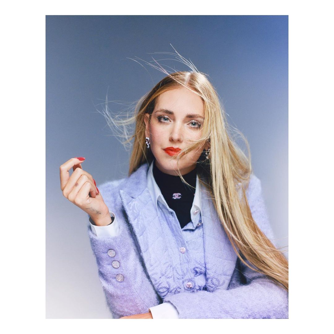 Adult woman with long blonde hair, wearing a light purple blazer with an embellished lapel against a grey studio backdrop.