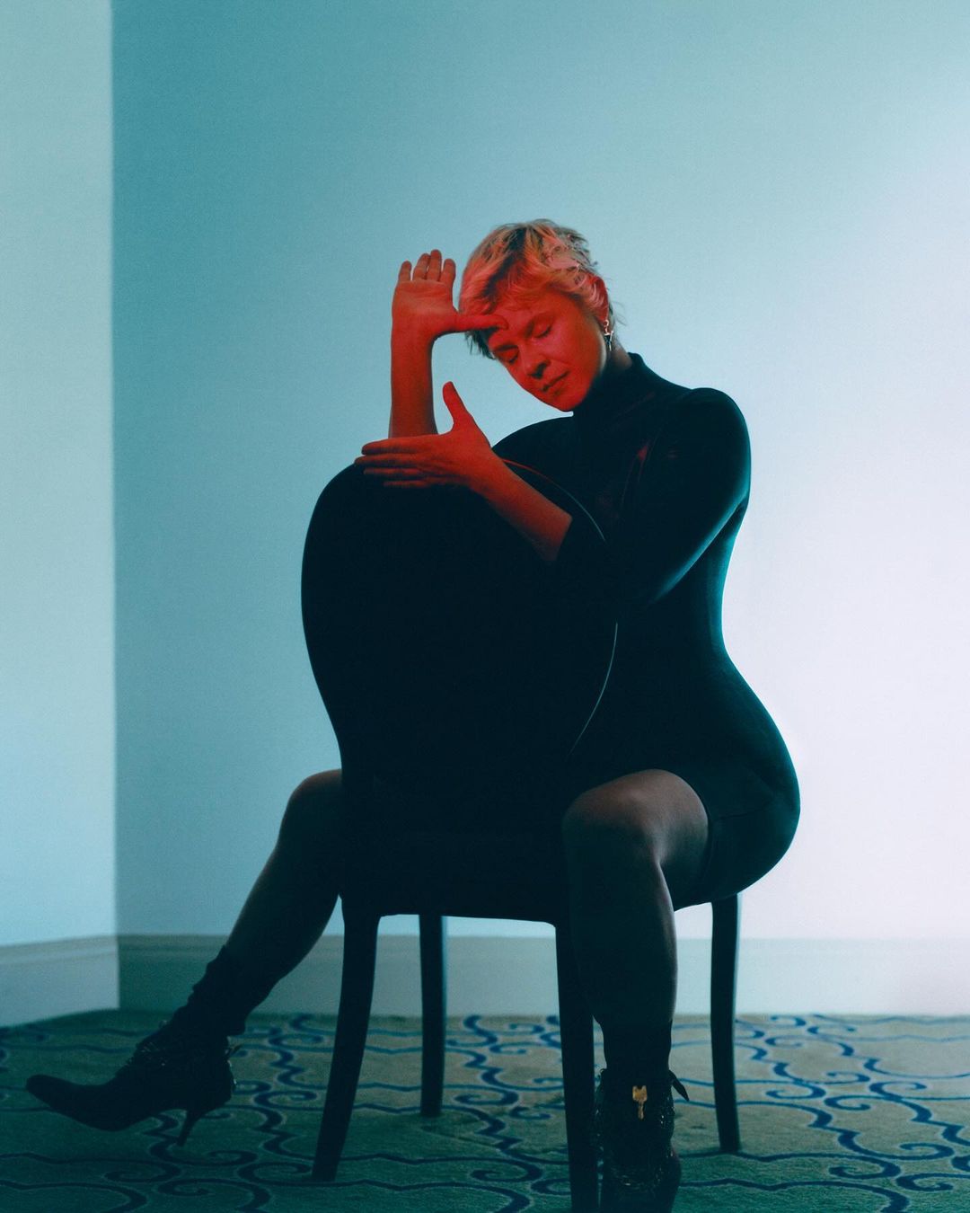 Woman with short blonde hair poses while sitting backwards on a chair in a studio with red and blue lighting effects.