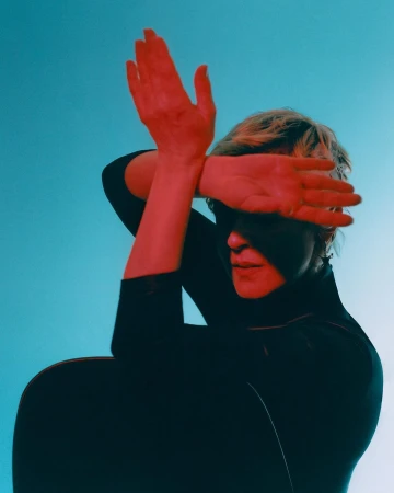 Woman in black turtleneck poses indoors with red lighting, face partially obscured by hand, against gradient blue background.