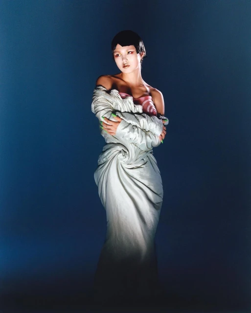 Asian woman in draped white couture dress and green nail polish poses against a dark blue backdrop under dramatic studio lighting.