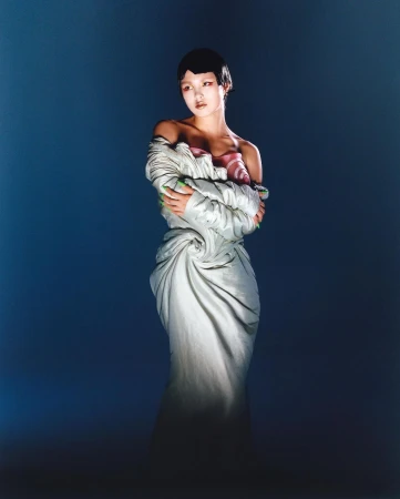 Asian woman in draped white couture dress and green nail polish poses against a dark blue backdrop under dramatic studio lighting.