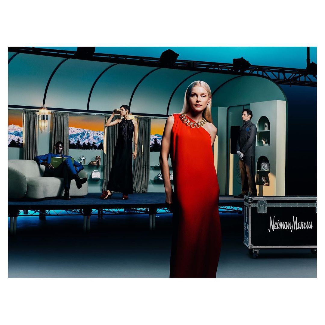 Fashion editorial shot indoors with a blonde model in a red dress, an African American model on a sofa, and a woman in a black dress.