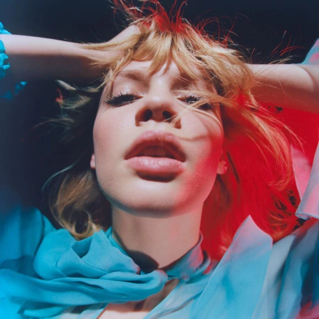 Eye-level close-up of a fair-skinned young adult female with blonde tousled hair. She wears a blue shawl, and colored gels accent the dark backdrop.