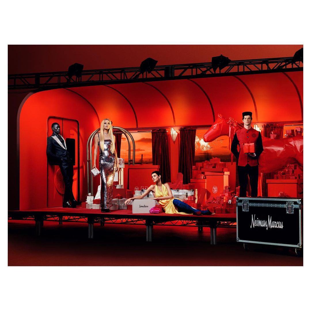 A red-toned fashion shoot features models on a stage, surrounded by a red set design and gift boxes in an indoor setting.