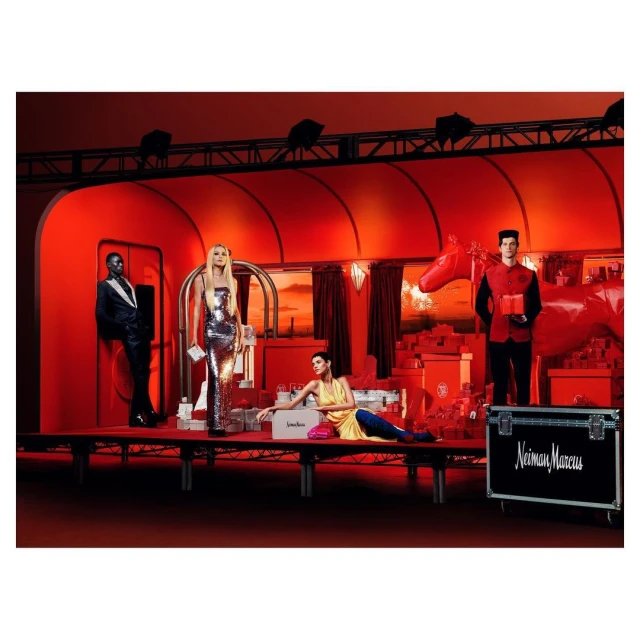 A red-toned fashion shoot features models on a stage, surrounded by a red set design and gift boxes in an indoor setting.