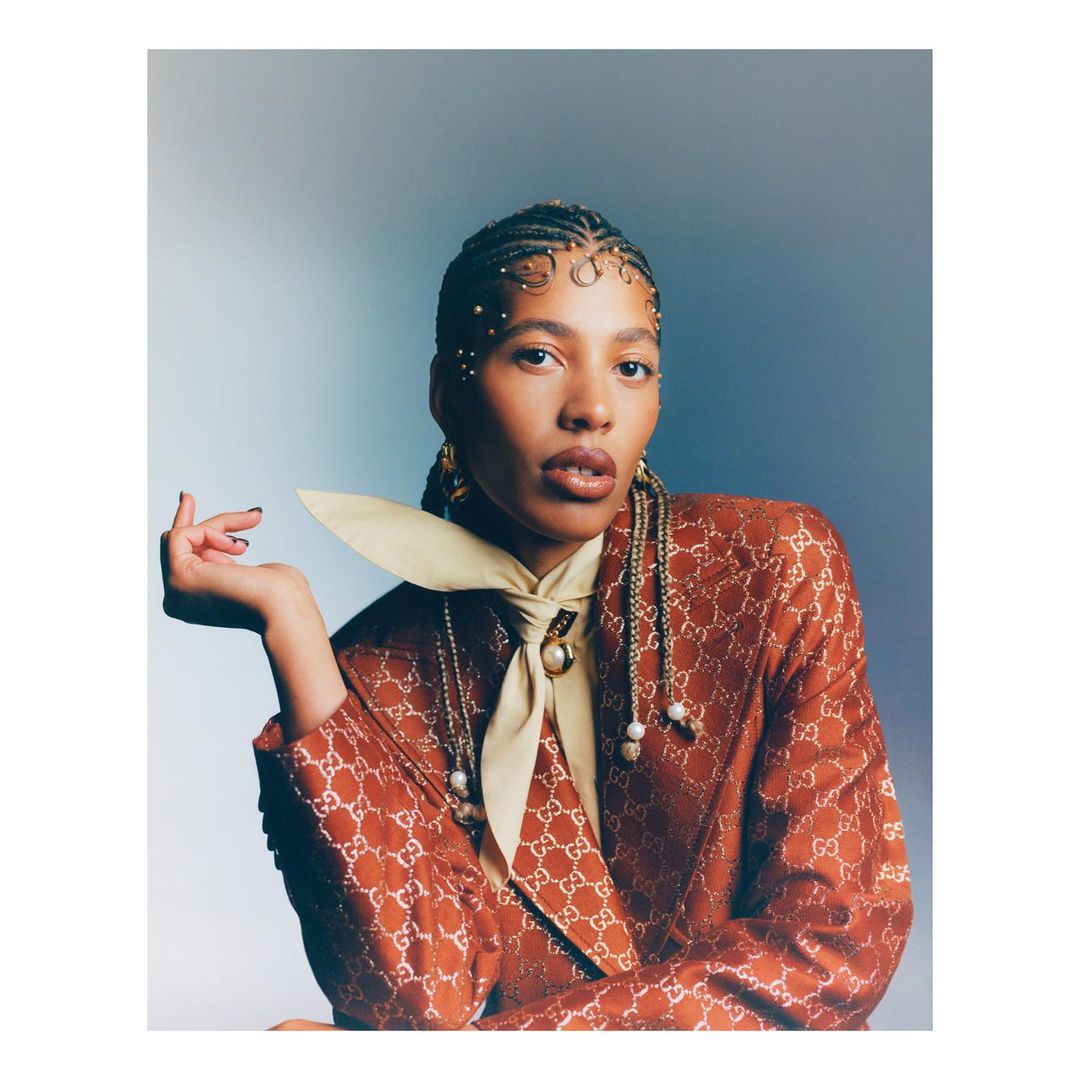 Portrait of an adult woman with braided hair, wearing a Gucci patterned jacket and tie against a gradient blue and white background.