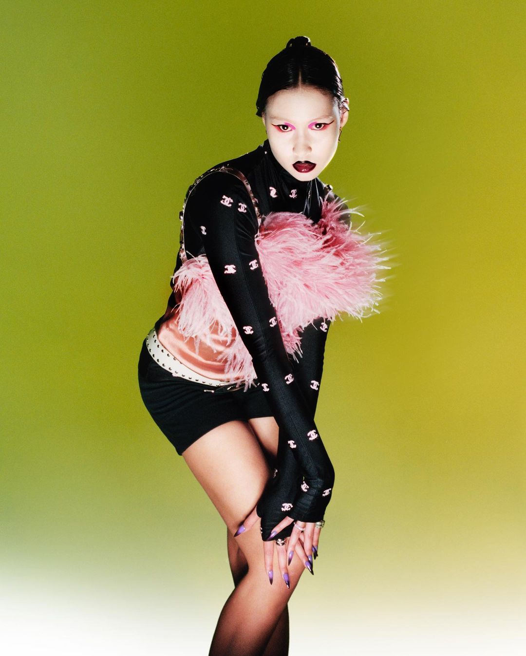 Young woman poses in a studio, wearing black top and skirt, pink feather boa, and high-fashion makeup against a yellow-green background.