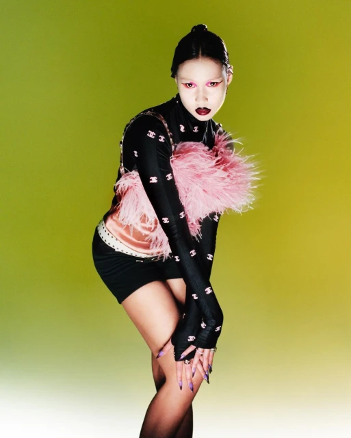 Young woman poses in a studio, wearing black top and skirt, pink feather boa, and high-fashion makeup against a yellow-green background.