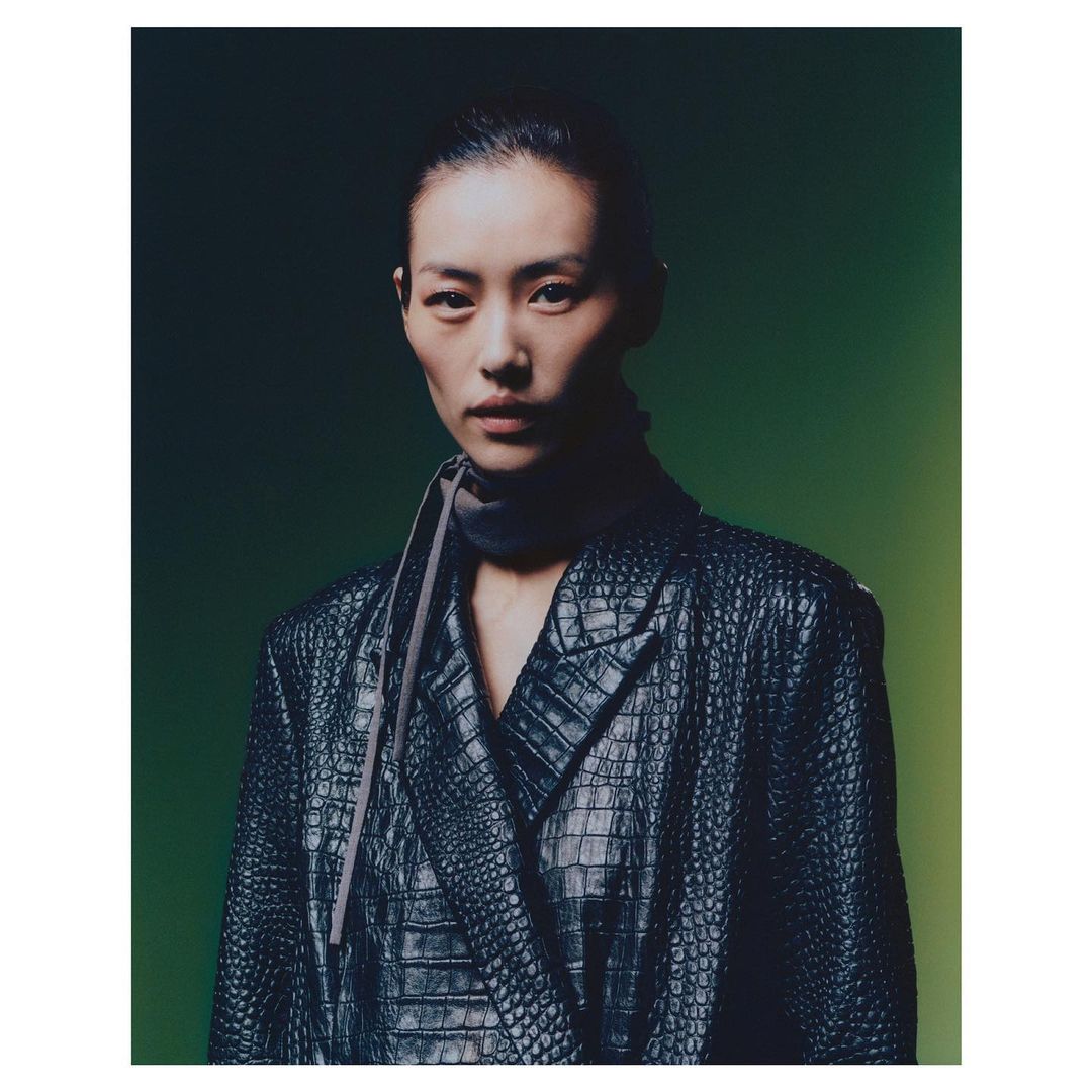 Portrait of an Asian woman in a reptile-textured black blazer, with a dark gray scarf, against a gradient green background.