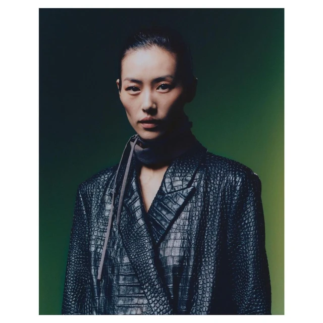 Portrait of an Asian woman in a reptile-textured black blazer, with a dark gray scarf, against a gradient green background.